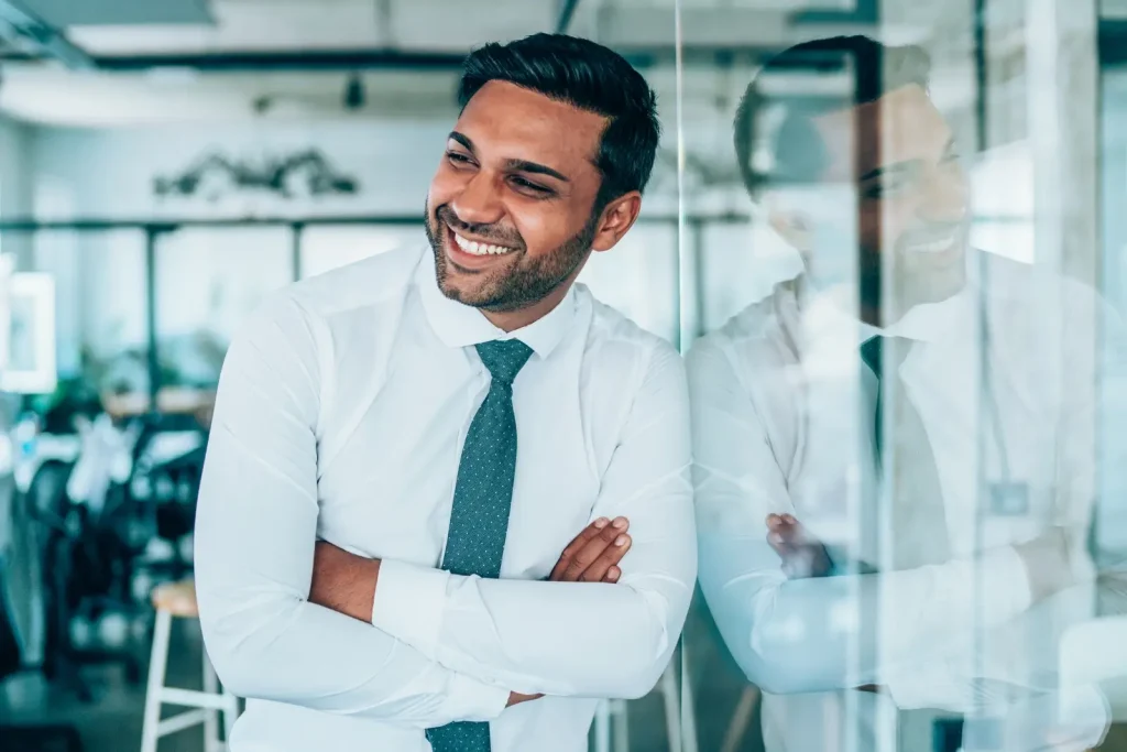 Smiling male financial advisor standing with arms crossed, reflected in a glass wall.