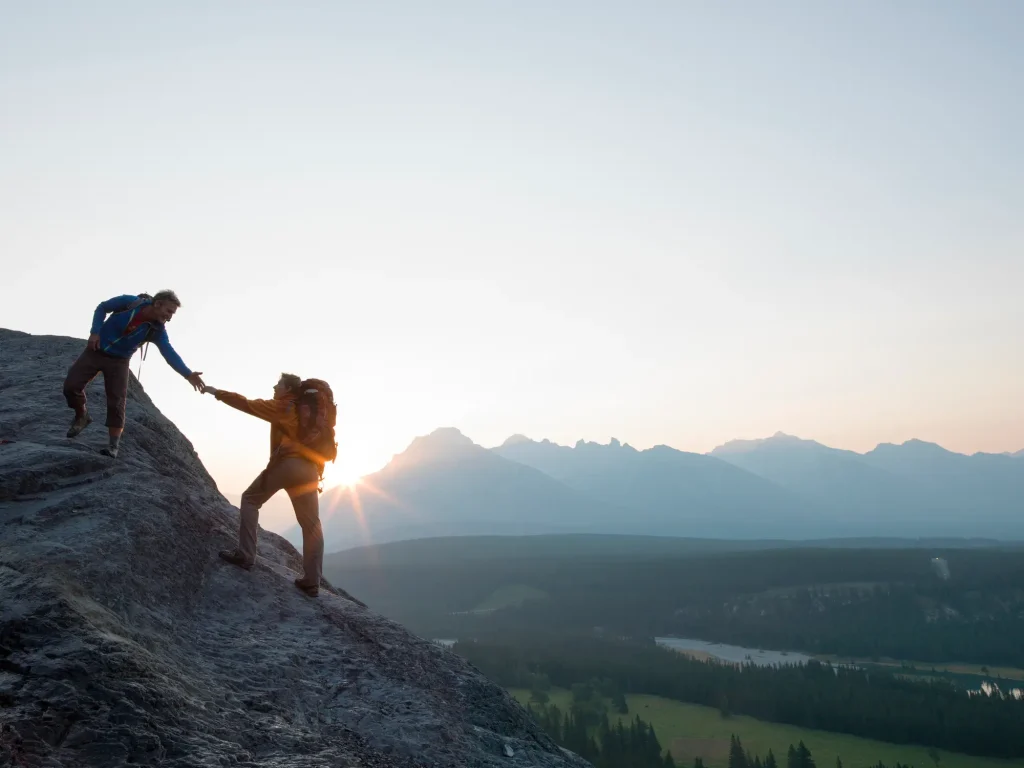 Two hikers on a mountain slope, one helping the other at sunrise.